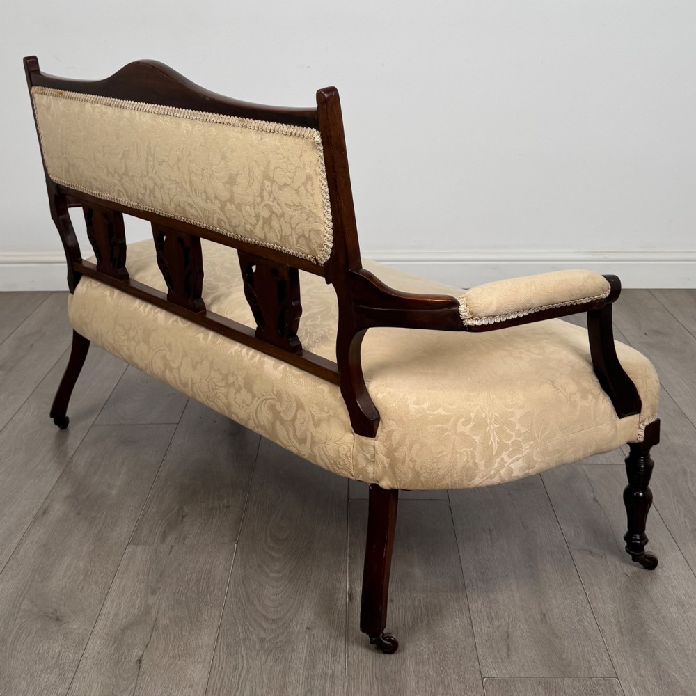 An antique Victorian rosewood settee featuring central backrest marquetry inlay, open armrests, turned front legs on castors, and cream damask upholstery. View VI