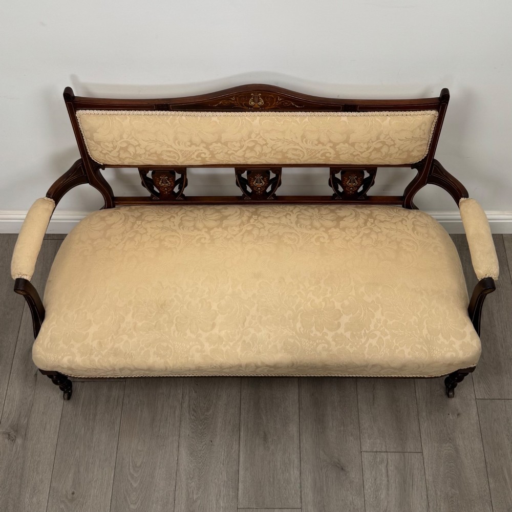 An antique Victorian rosewood settee featuring central backrest marquetry inlay, open armrests, turned front legs on castors, and cream damask upholstery. View IX