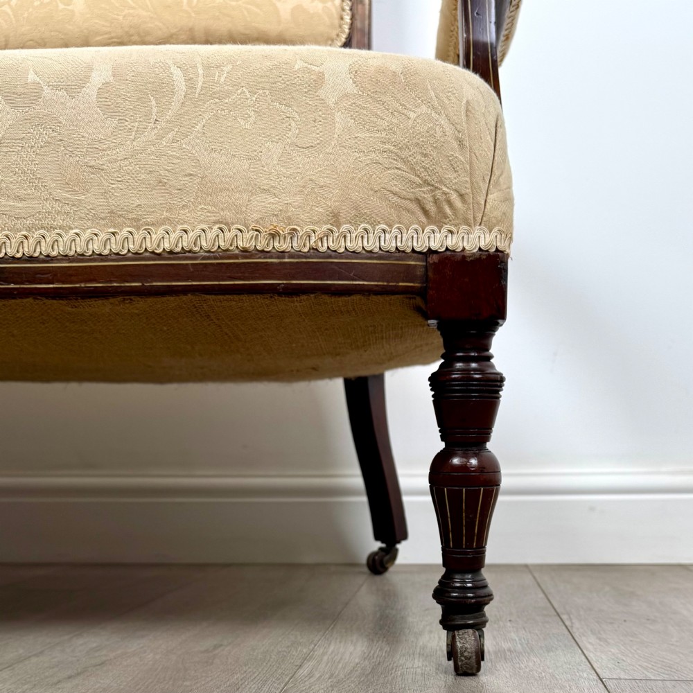 An antique Victorian rosewood settee featuring central backrest marquetry inlay, open armrests, turned front legs on castors, and cream damask upholstery. View X
