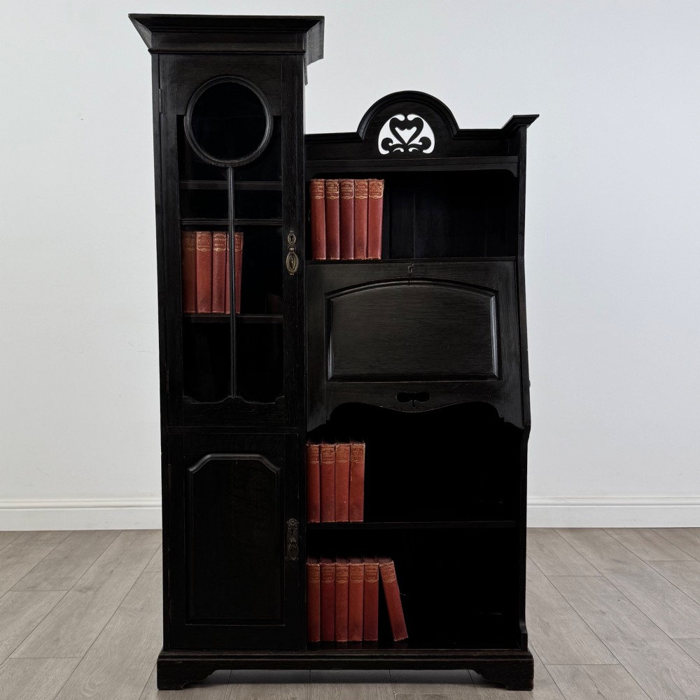 An ebonised oak bureau bookcase featuring an asymmetrical design with a glazed door cabinet, a slanted fall-front writing surface, open shelving, and a heart cutout crest.