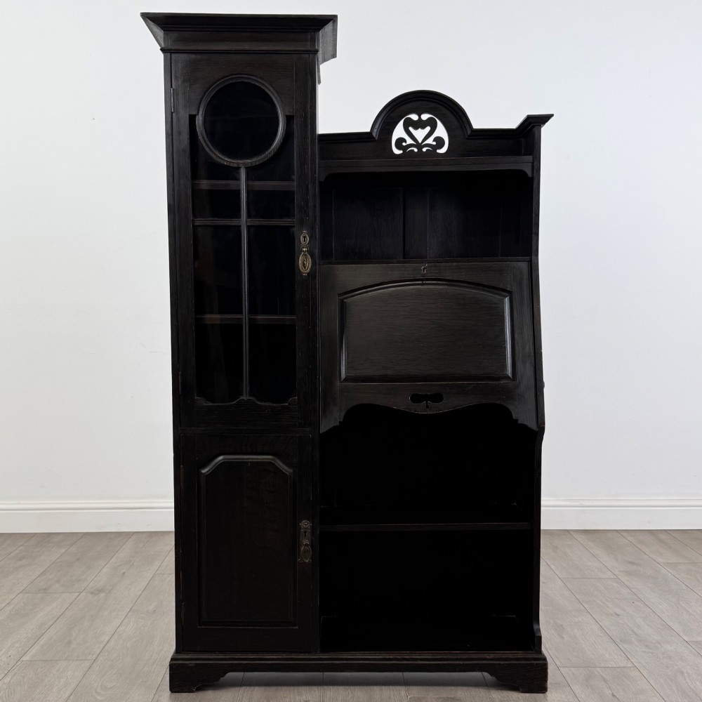 An ebonised oak bureau bookcase featuring an asymmetrical design with a glazed door cabinet, a slanted fall-front writing surface, open shelving, and a heart cutout crest. View II