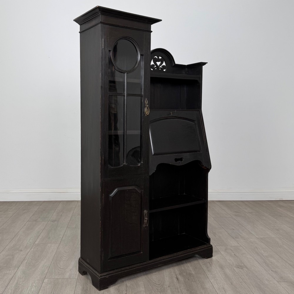 An ebonised oak bureau bookcase featuring an asymmetrical design with a glazed door cabinet, a slanted fall-front writing surface, open shelving, and a heart cutout crest. View X