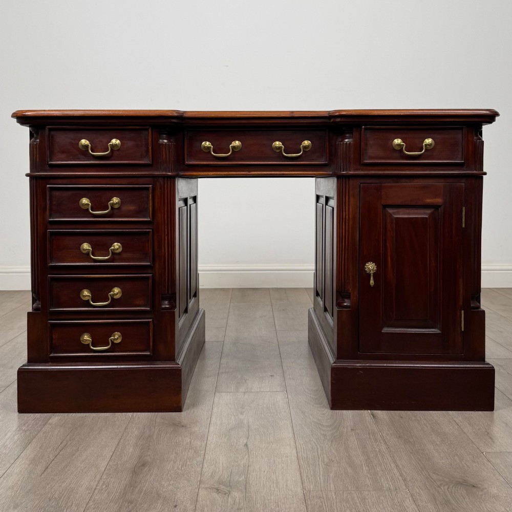 A dark mahogany pedestal desk featuring a central kneehole drawer flanked by five drawers on the left and a panelled cupboard door on the right, all fitted with brass drop handles.
