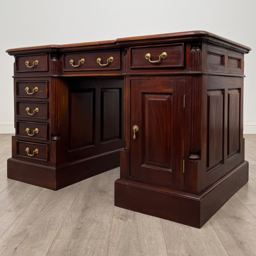 A dark mahogany pedestal desk featuring a central kneehole drawer flanked by five drawers on the left and a panelled cupboard door on the right, all fitted with brass drop handles. View III