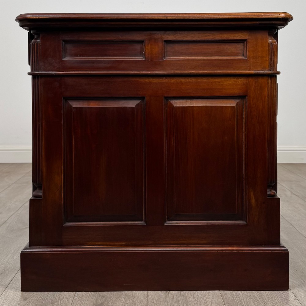 A dark mahogany pedestal desk featuring a central kneehole drawer flanked by five drawers on the left and a panelled cupboard door on the right, all fitted with brass drop handles. View IV