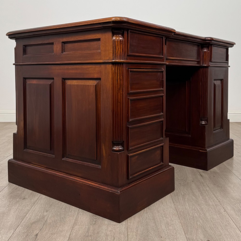 A dark mahogany pedestal desk featuring a central kneehole drawer flanked by five drawers on the left and a panelled cupboard door on the right, all fitted with brass drop handles. View V