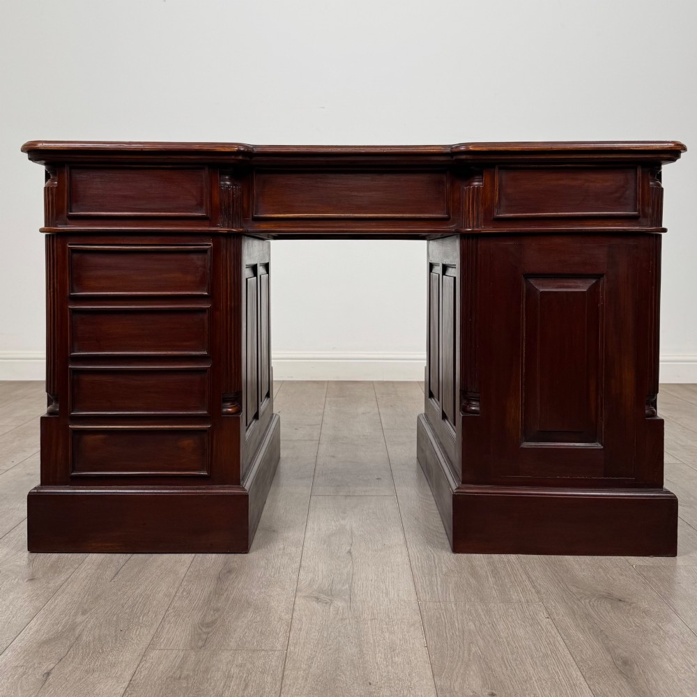 A dark mahogany pedestal desk featuring a central kneehole drawer flanked by five drawers on the left and a panelled cupboard door on the right, all fitted with brass drop handles. View VI