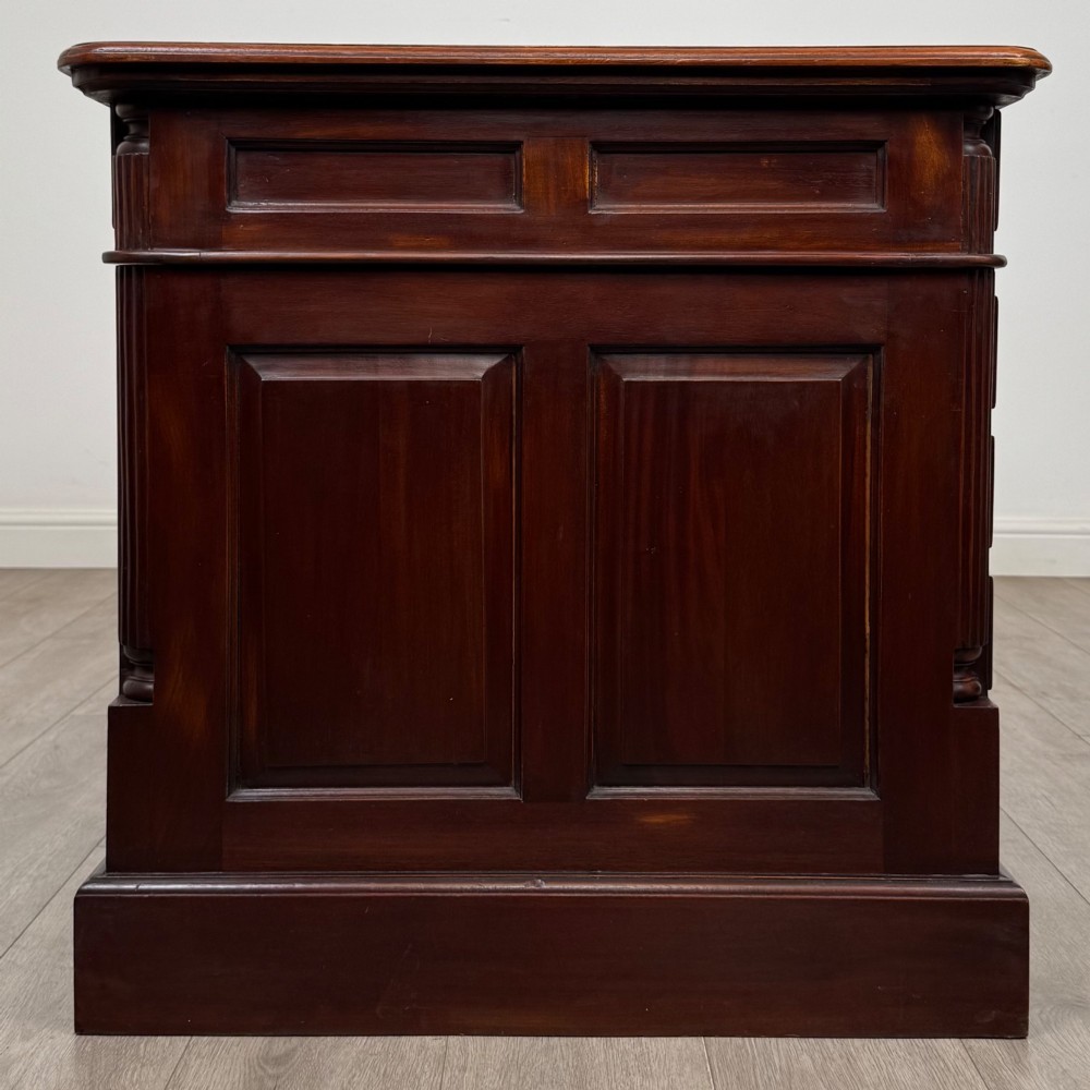 A dark mahogany pedestal desk featuring a central kneehole drawer flanked by five drawers on the left and a panelled cupboard door on the right, all fitted with brass drop handles. View VIII