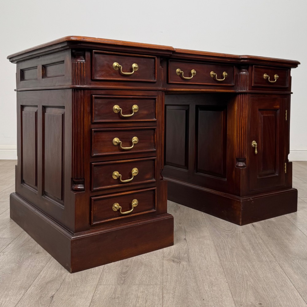 A dark mahogany pedestal desk featuring a central kneehole drawer flanked by five drawers on the left and a panelled cupboard door on the right, all fitted with brass drop handles. View IX