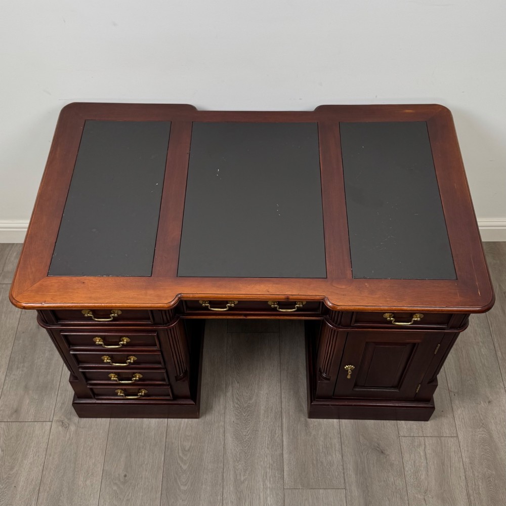 A dark mahogany pedestal desk featuring a central kneehole drawer flanked by five drawers on the left and a panelled cupboard door on the right, all fitted with brass drop handles. View X