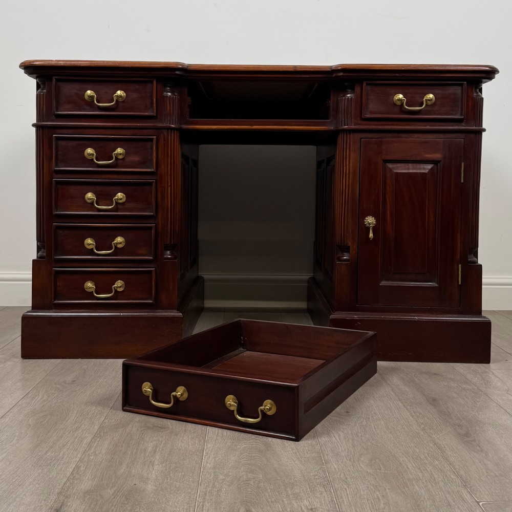 A dark mahogany pedestal desk featuring a central kneehole drawer flanked by five drawers on the left and a panelled cupboard door on the right, all fitted with brass drop handles. View XI