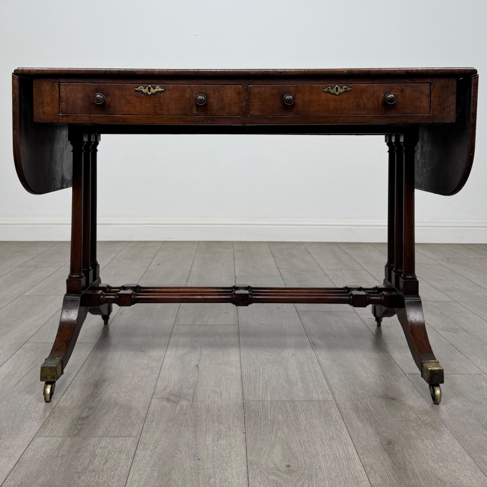 A 19th-century Regency style yew wood side table featuring drop-leaf ends, twin drawers with brass escutcheons and round knobs, turned twin-stem supports, a stretcher, and brass castors.