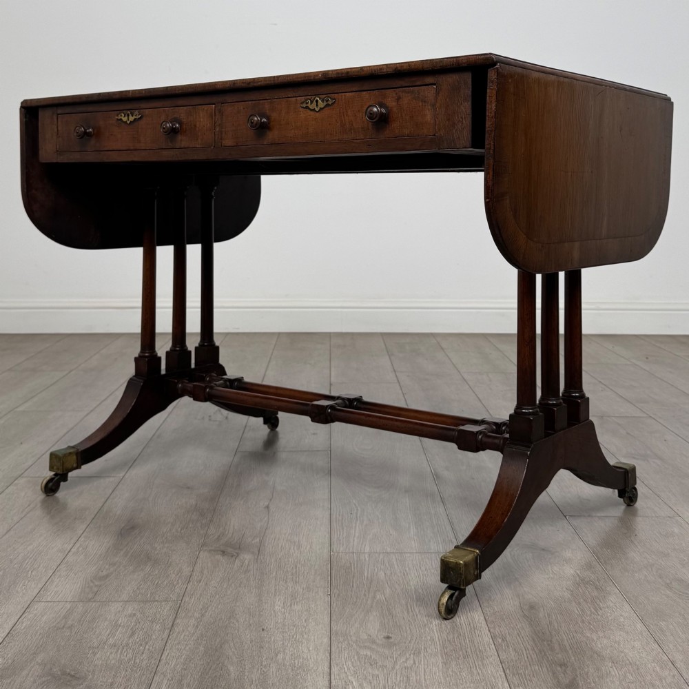 A 19th-century Regency style yew wood side table featuring drop-leaf ends, twin drawers with brass escutcheons and round knobs, turned twin-stem supports, a stretcher, and brass castors. View III