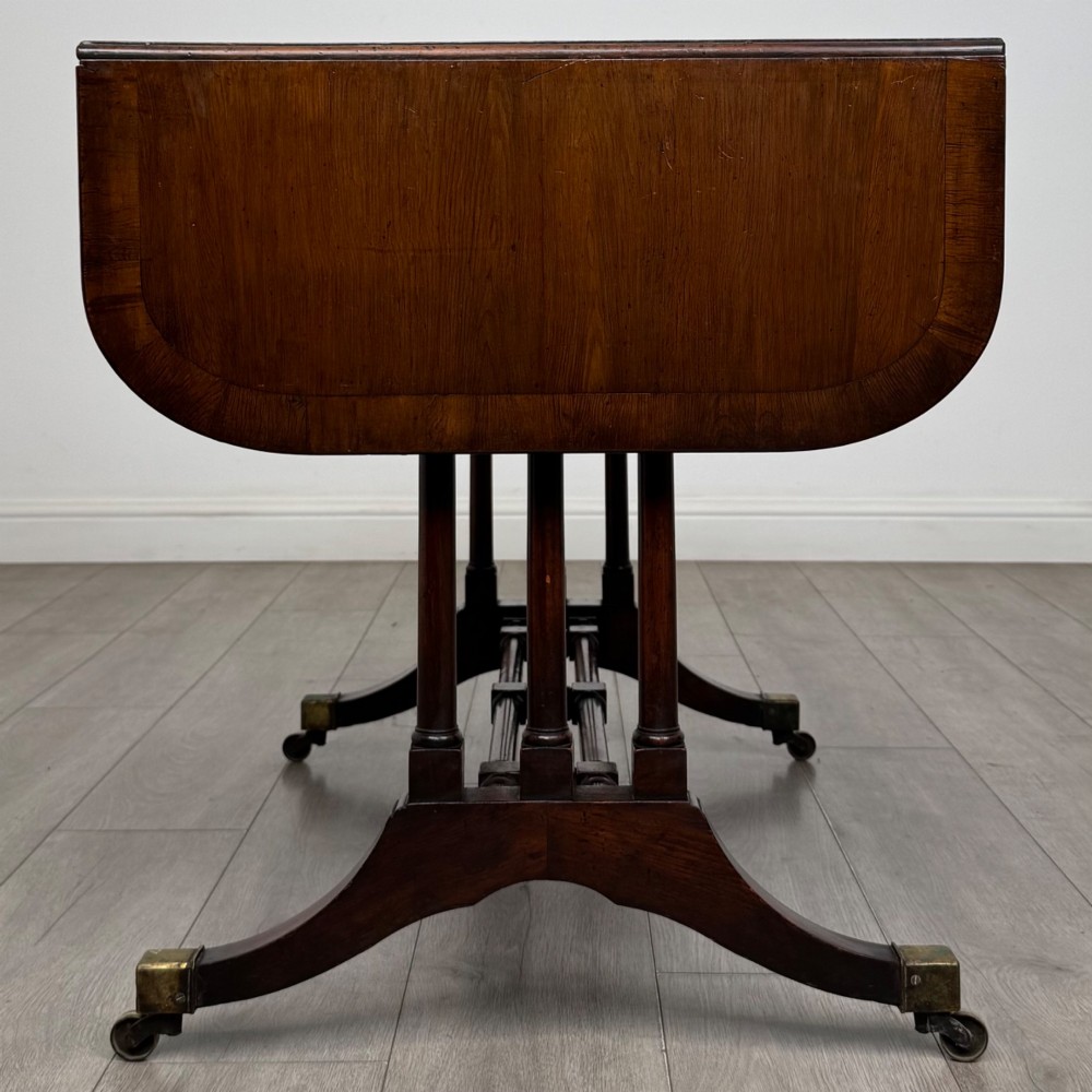 A 19th-century Regency style yew wood side table featuring drop-leaf ends, twin drawers with brass escutcheons and round knobs, turned twin-stem supports, a stretcher, and brass castors. View IV