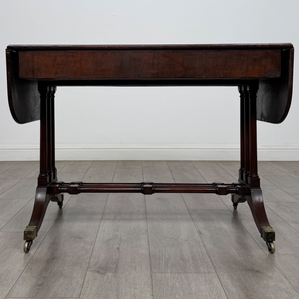 A 19th-century Regency style yew wood side table featuring drop-leaf ends, twin drawers with brass escutcheons and round knobs, turned twin-stem supports, a stretcher, and brass castors. View VI