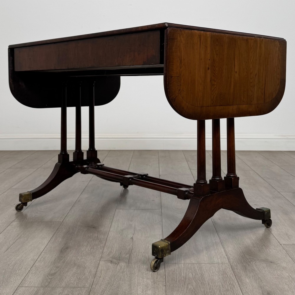 A 19th-century Regency style yew wood side table featuring drop-leaf ends, twin drawers with brass escutcheons and round knobs, turned twin-stem supports, a stretcher, and brass castors. View VII