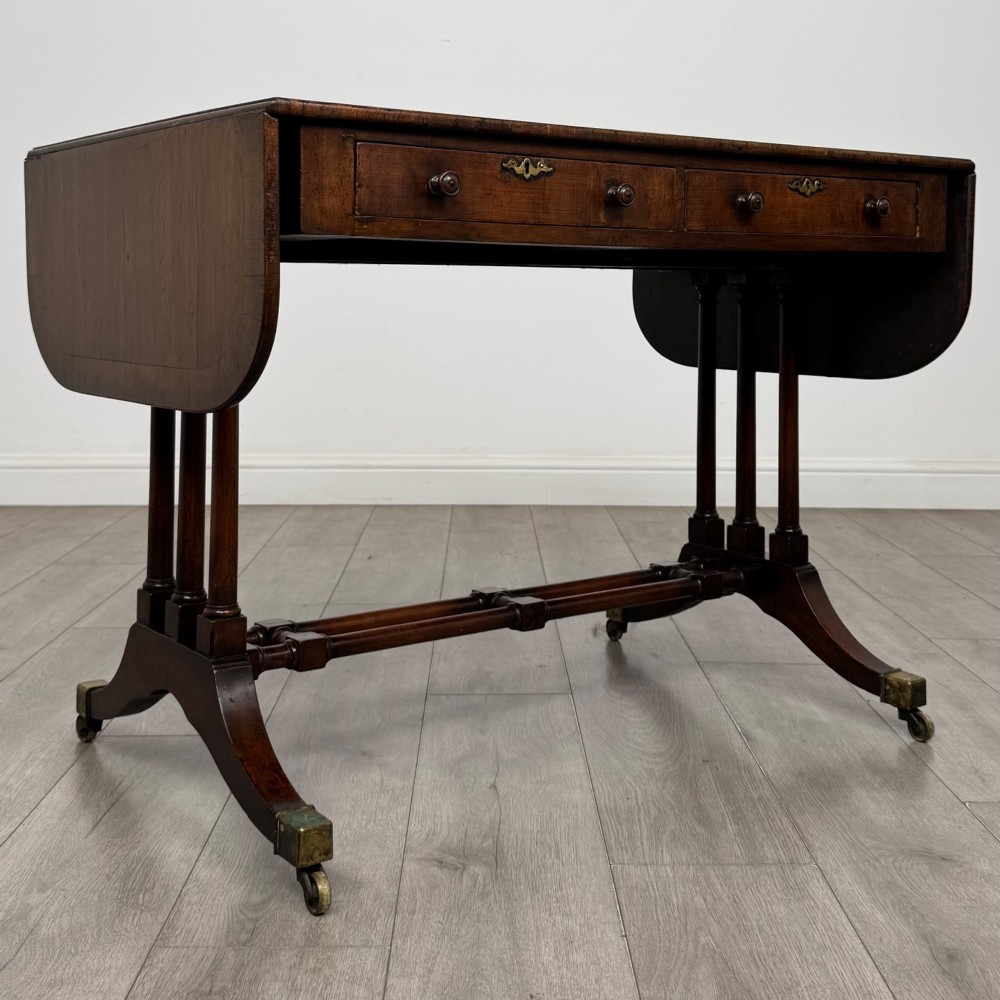 A 19th-century Regency style yew wood side table featuring drop-leaf ends, twin drawers with brass escutcheons and round knobs, turned twin-stem supports, a stretcher, and brass castors. View IX