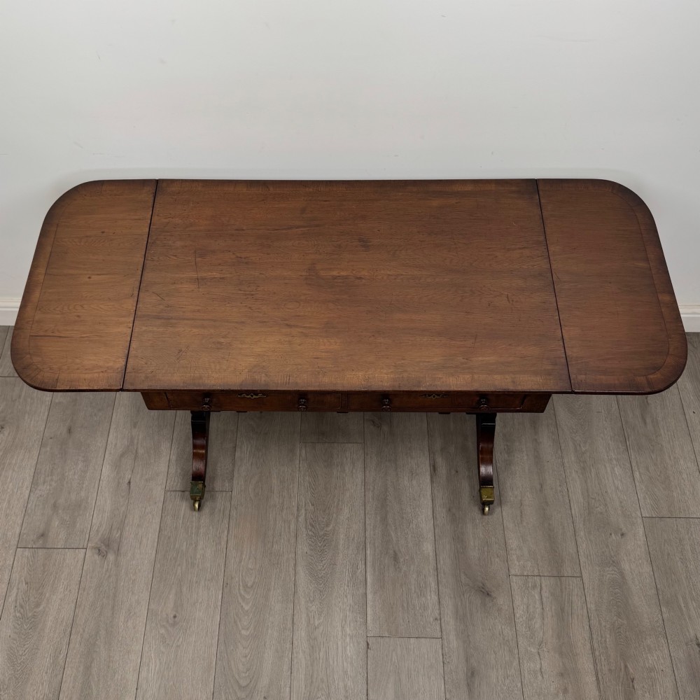 A 19th-century Regency style yew wood side table featuring drop-leaf ends, twin drawers with brass escutcheons and round knobs, turned twin-stem supports, a stretcher, and brass castors. View XI