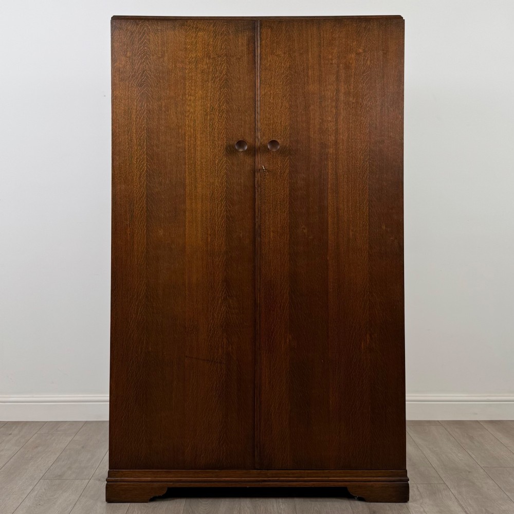 A freestanding mid-20th-century double wardrobe crafted from medium brown oak with twin flush doors, small circular wooden handles, and a low plinth base.