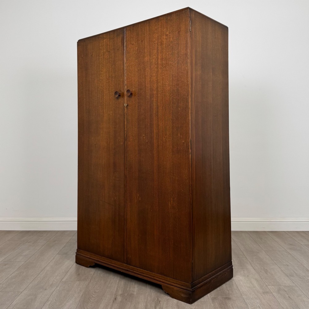 A freestanding mid-20th-century double wardrobe crafted from medium brown oak with twin flush doors, small circular wooden handles, and a low plinth base. View III