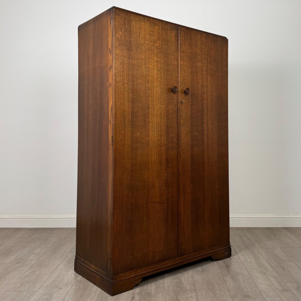 A freestanding mid-20th-century double wardrobe crafted from medium brown oak with twin flush doors, small circular wooden handles, and a low plinth base. View IX