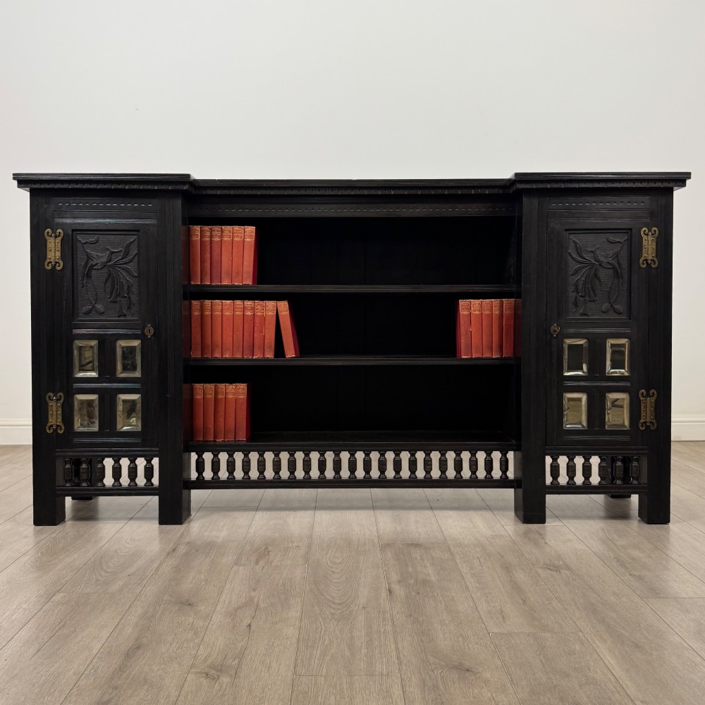 An antique ebonised black wood sideboard featuring a central open bookshelf, two carved side doors with glass panels, brass hardware, and a spindled lower rail.