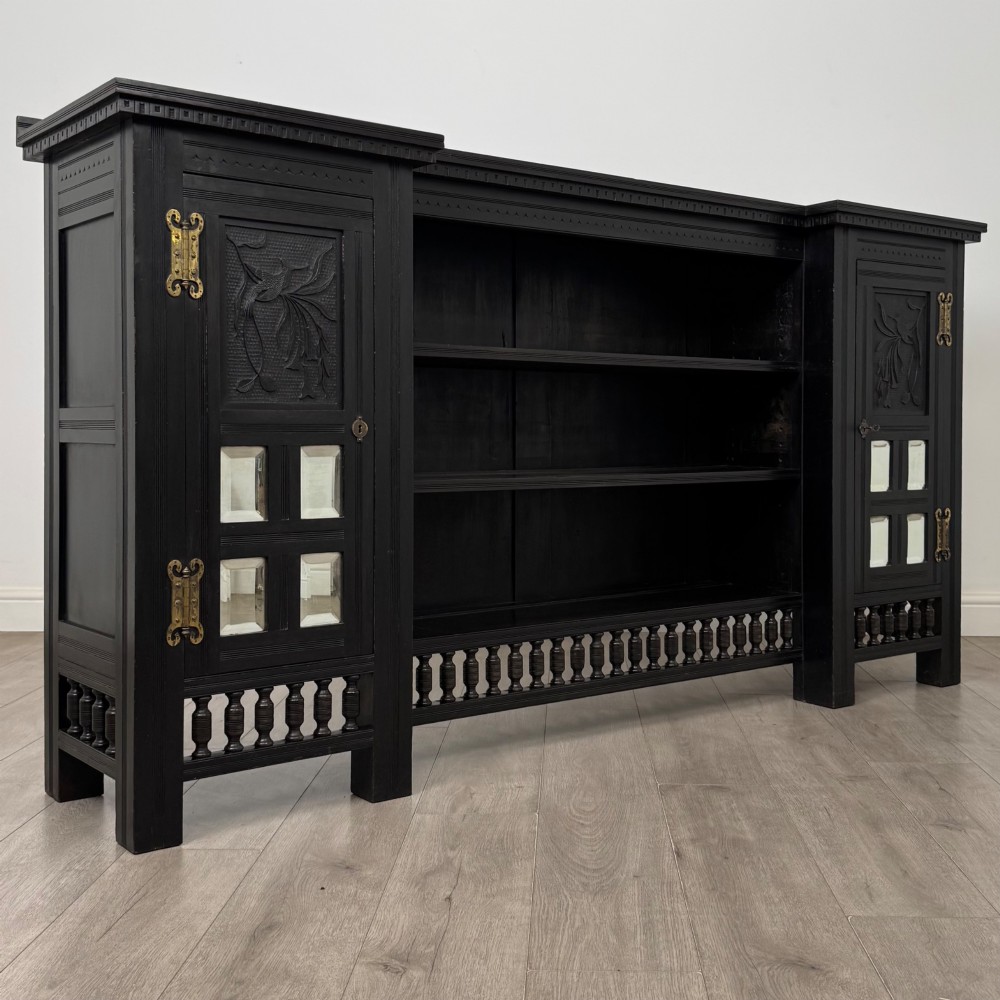 An antique ebonised black wood sideboard featuring a central open bookshelf, two carved side doors with glass panels, brass hardware, and a spindled lower rail. View X