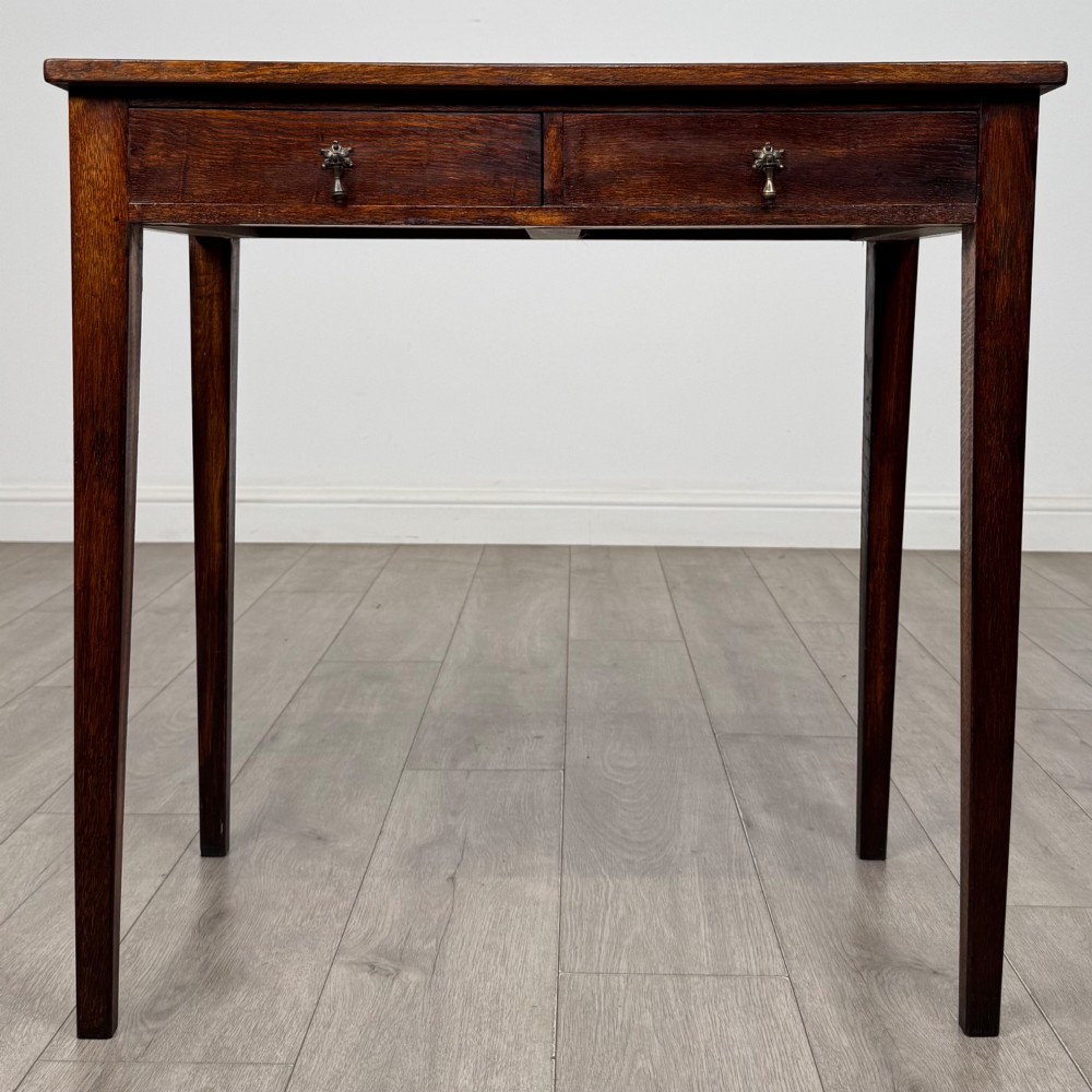 An antique dark oak Edwardian side table with two front drawers, metallic drop handles, and straight tapered legs.
