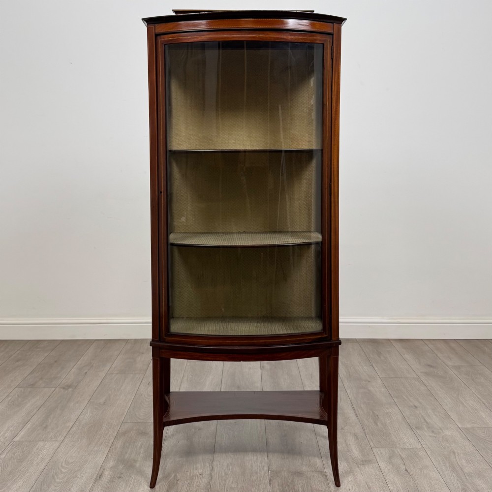 An Edwardian mahogany display cabinet with a bow-front glazed door, fabric-lined interior shelves, marquetry inlay, and slender tapered legs with a lower shelf.