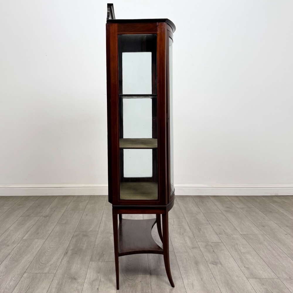 An Edwardian mahogany display cabinet with a bow-front glazed door, fabric-lined interior shelves, marquetry inlay, and slender tapered legs with a lower shelf. View VIII