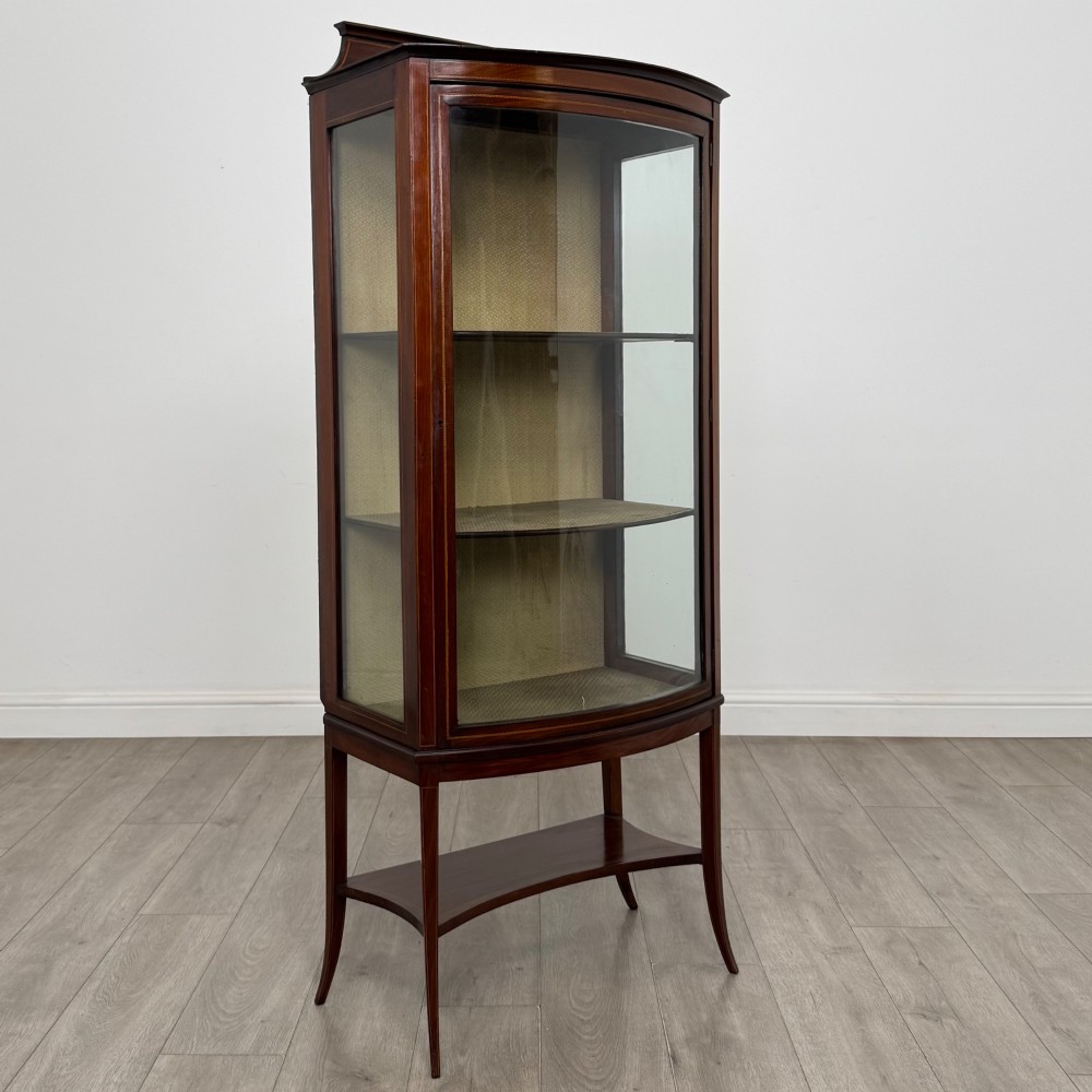 An Edwardian mahogany display cabinet with a bow-front glazed door, fabric-lined interior shelves, marquetry inlay, and slender tapered legs with a lower shelf. View IX