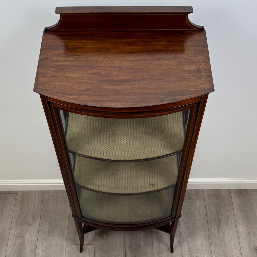 An Edwardian mahogany display cabinet with a bow-front glazed door, fabric-lined interior shelves, marquetry inlay, and slender tapered legs with a lower shelf. View X