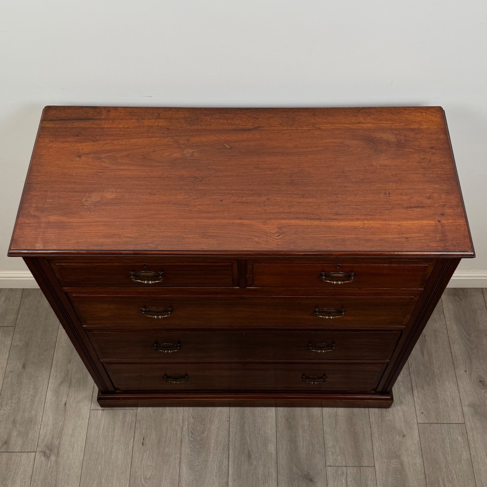 An Edwardian walnut chest of drawers featuring a two over three drawer layout with brass drop handles and a solid plinth base. View IX
