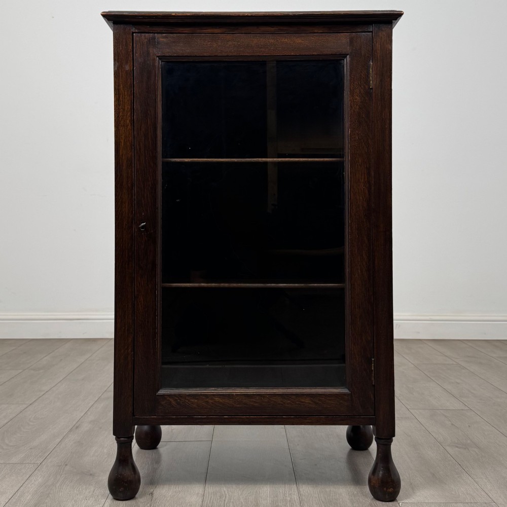 An Edwardian dark brown oak display cabinet featuring a single glazed door and turned feet.
