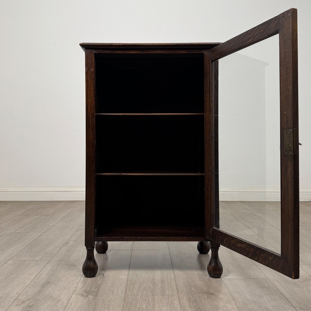 An Edwardian dark brown oak display cabinet featuring a single glazed door and turned feet. View II