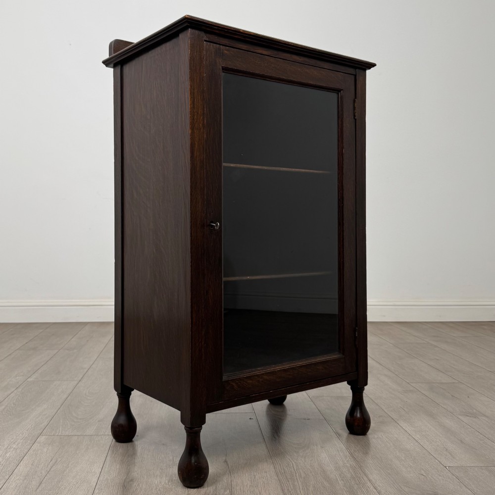 An Edwardian dark brown oak display cabinet featuring a single glazed door and turned feet. View IX