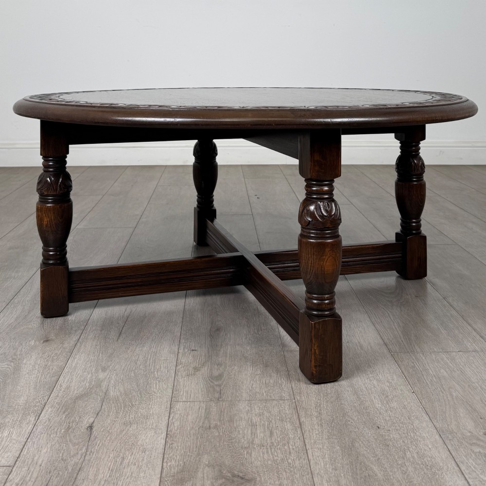 A low circular dark oak coffee table featuring carved turned legs, an X-shaped cross stretcher, and a carved geometric border along the top rim. View II