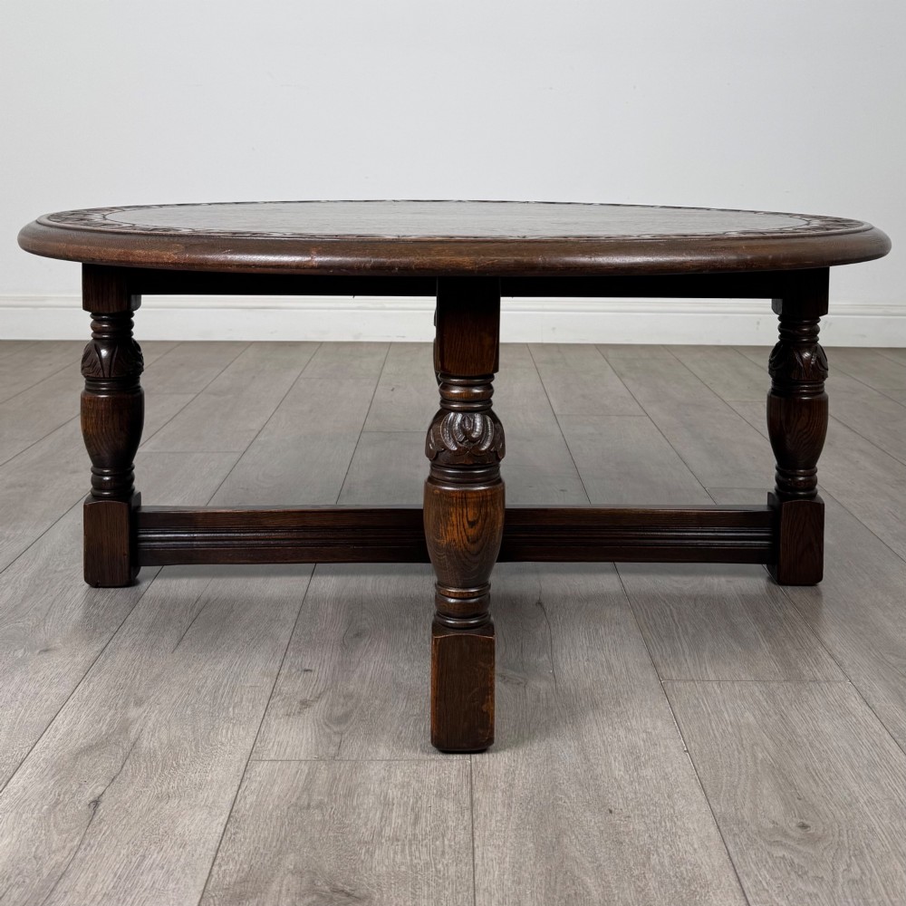A low circular dark oak coffee table featuring carved turned legs, an X-shaped cross stretcher, and a carved geometric border along the top rim. View V