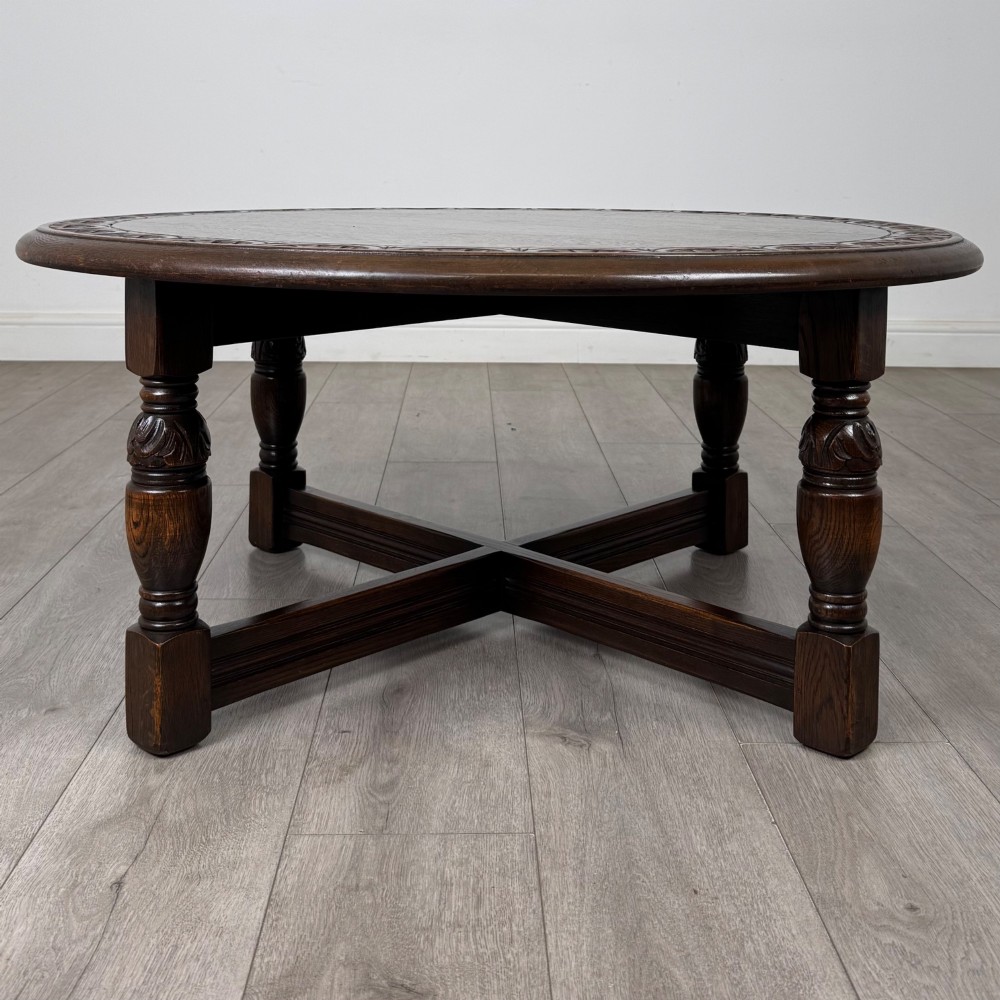 A low circular dark oak coffee table featuring carved turned legs, an X-shaped cross stretcher, and a carved geometric border along the top rim. View VI