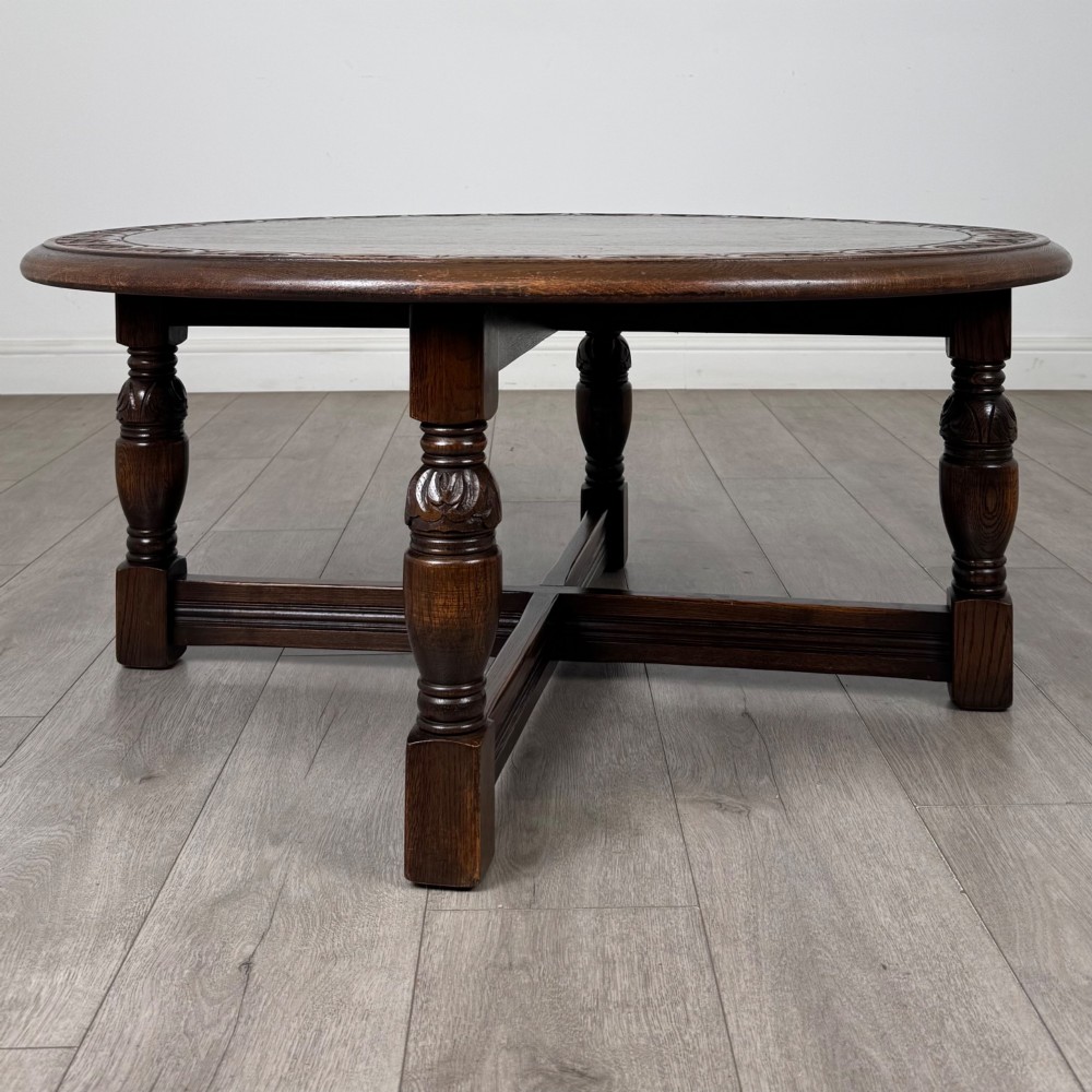 A low circular dark oak coffee table featuring carved turned legs, an X-shaped cross stretcher, and a carved geometric border along the top rim. View VII
