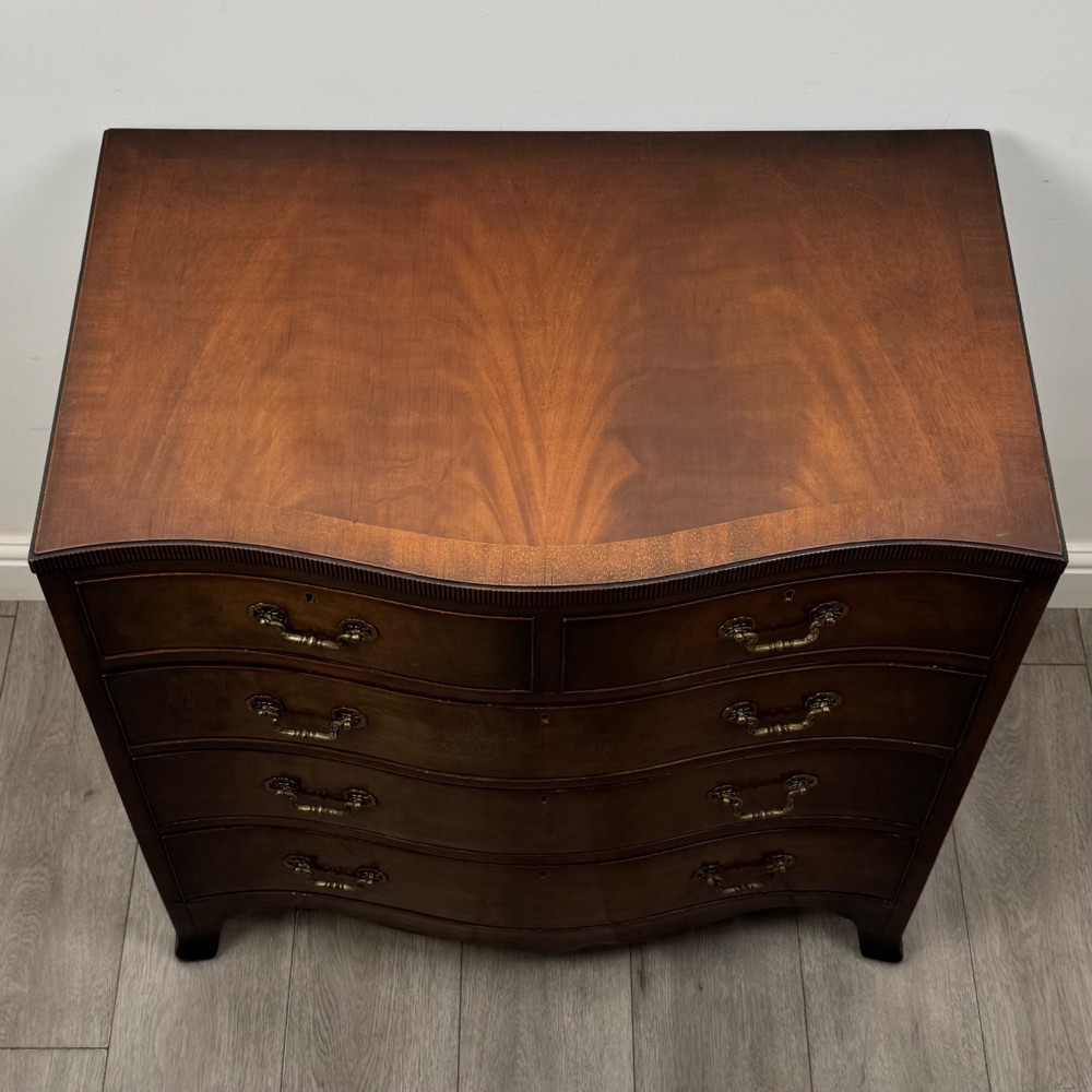 A mahogany serpentine chest of drawers featuring four graduating drawer tiers with brass handles and a curved front profile. View IX