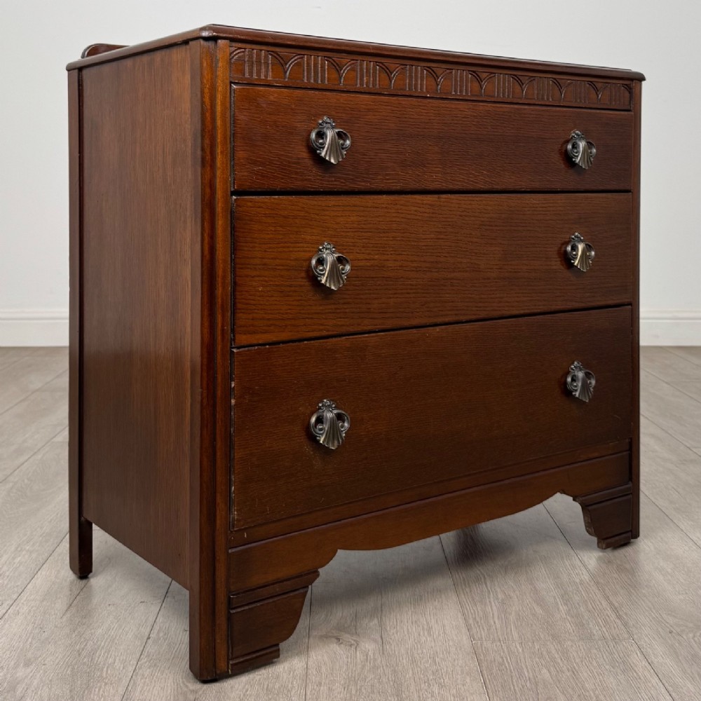 A dark brown oak chest of drawers featuring three wide drawers with ornamental metal drop handles and a carved geometric frieze along the top edge. View VIII