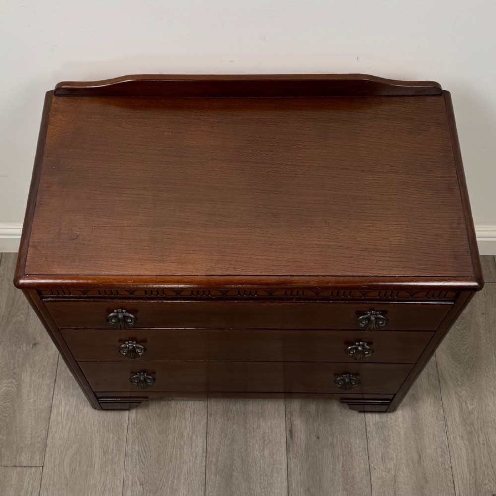A dark brown oak chest of drawers featuring three wide drawers with ornamental metal drop handles and a carved geometric frieze along the top edge. View IX