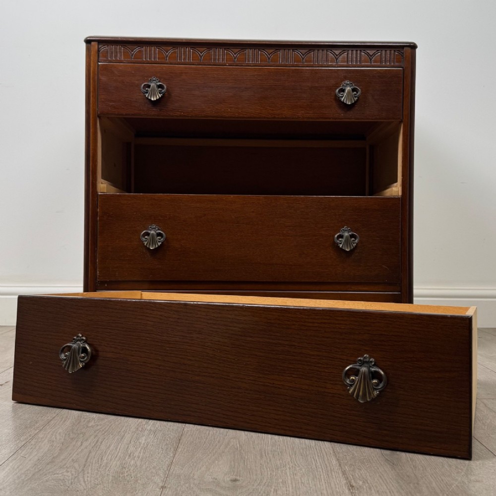 A dark brown oak chest of drawers featuring three wide drawers with ornamental metal drop handles and a carved geometric frieze along the top edge. View X