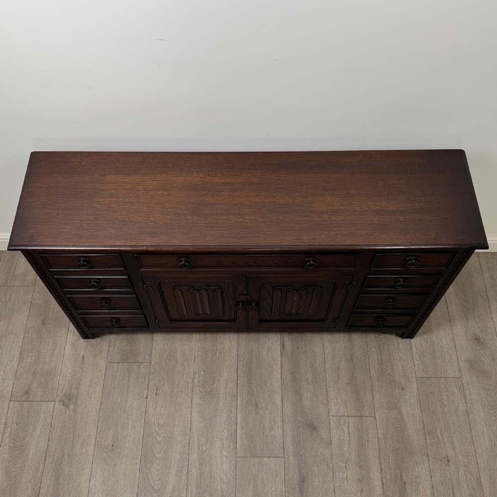 A dark oak sideboard featuring carved linenfold central doors flanked by eight drawers with metal drop ring handles. View X