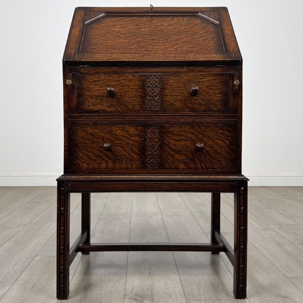 A small Edwardian dark oak writing bureau on a tall stand with two drawers featuring carved central panels and turned knobs.