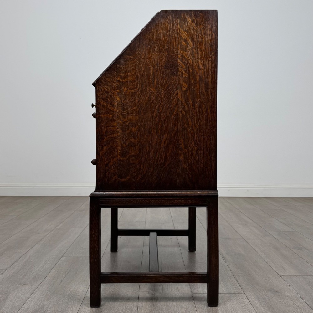 A small Edwardian dark oak writing bureau on a tall stand with two drawers featuring carved central panels and turned knobs. View IV
