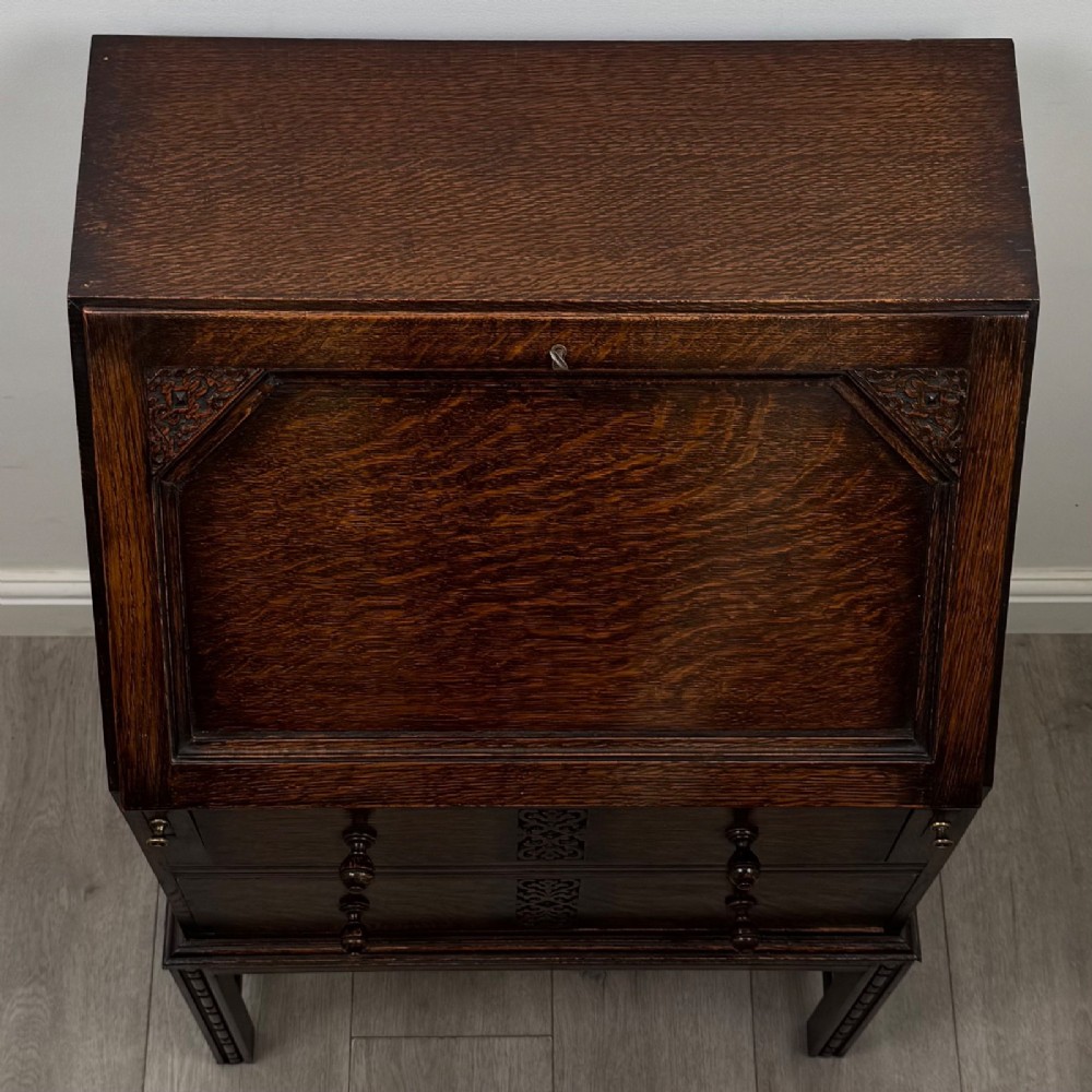 A small Edwardian dark oak writing bureau on a tall stand with two drawers featuring carved central panels and turned knobs. View X