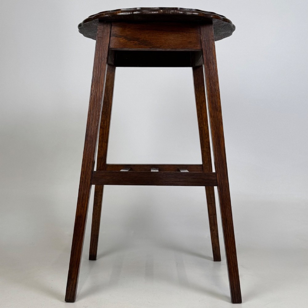 A tall, dark oak Edwardian side table featuring a scalloped piecrust rim top, four splayed square legs, and a lower slatted shelf.
