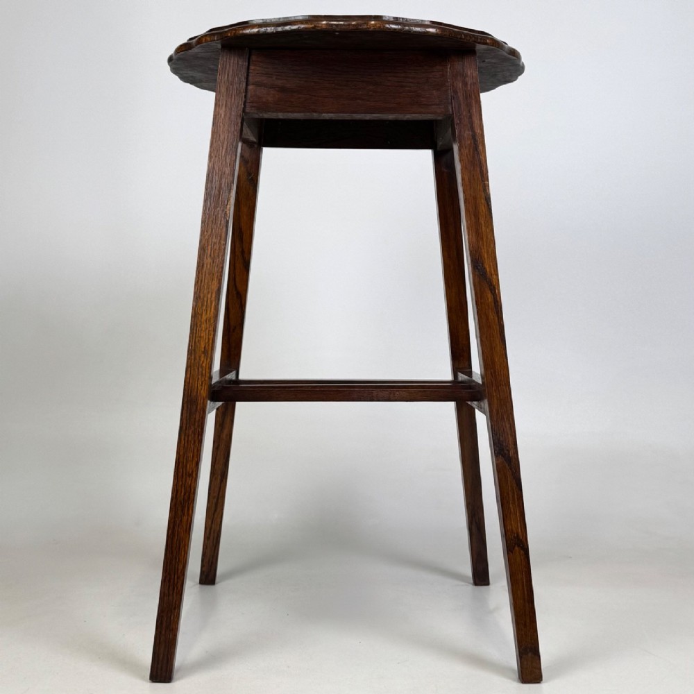 A tall, dark oak Edwardian side table featuring a scalloped piecrust rim top, four splayed square legs, and a lower slatted shelf. View III